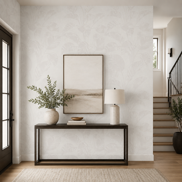 Erismann Opus 10459-14 warm neutral botanical leaf wallpaper in a bright entryway with a slim console table, framed artwork, ceramic lamp, greenery, staircase, and soft modern styling.