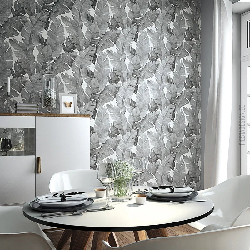 Marburg Avalon 31624 Grayscale tropical banana leaf wallpaper on a dining room wall, shown with a round table set with plates and glasses, white chairs, a sideboard, and a window with curtains.