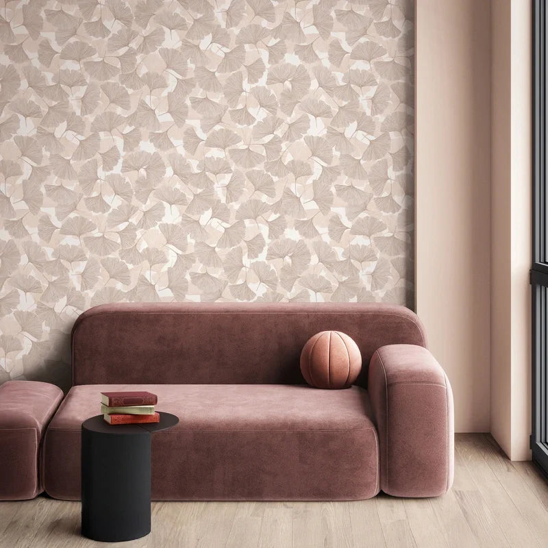 Marburg Boho 35780 Beige and light brown ginkgo leaf wallpaper behind a modern curved sofa, with a small black side table holding books, a round pillow on the sofa, and a window beside the scene.