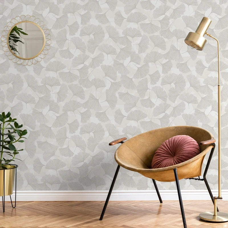 Marburg Boho 35782 Beige and grey ginkgo leaf wallpaper shown in a styled room with a tan lounge chair holding a round pink cushion, a gold floor lamp, a small round mirror, and a potted plant on a stand.
