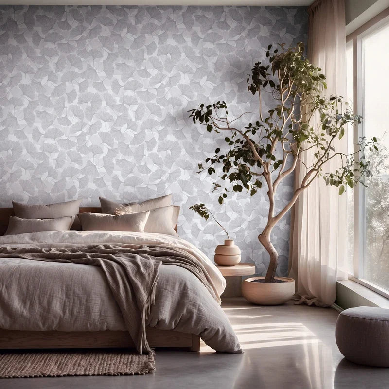 Marburg Boho 35783 Bedroom interior featuring a gray leaf-pattern wallpaper on the wall behind a low bed. A large potted tree stands by a window with sheer curtains, with neutral bedding and a rug in view.