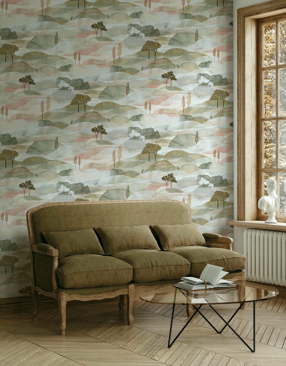 Marburg Boho 35785 Scenic watercolor hills wallpaper with small stylized trees in muted green and blush tones, shown behind a cushioned sofa, a glass coffee table with an open book, and a window with a white sculptural bust on the sill.