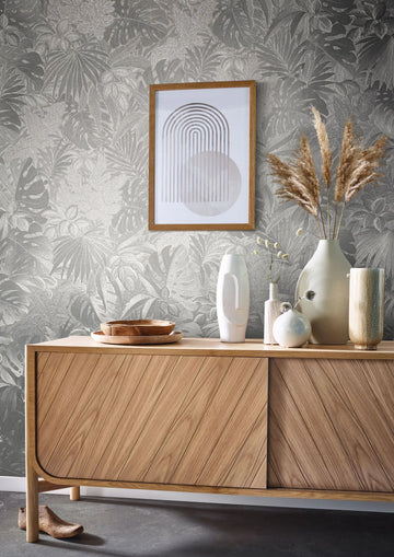 Marburg Botanica 33301 Silver botanical leaf wallpaper behind a wood sideboard styled with ceramic vases, pampas grass, wooden bowls, and a framed geometric print.