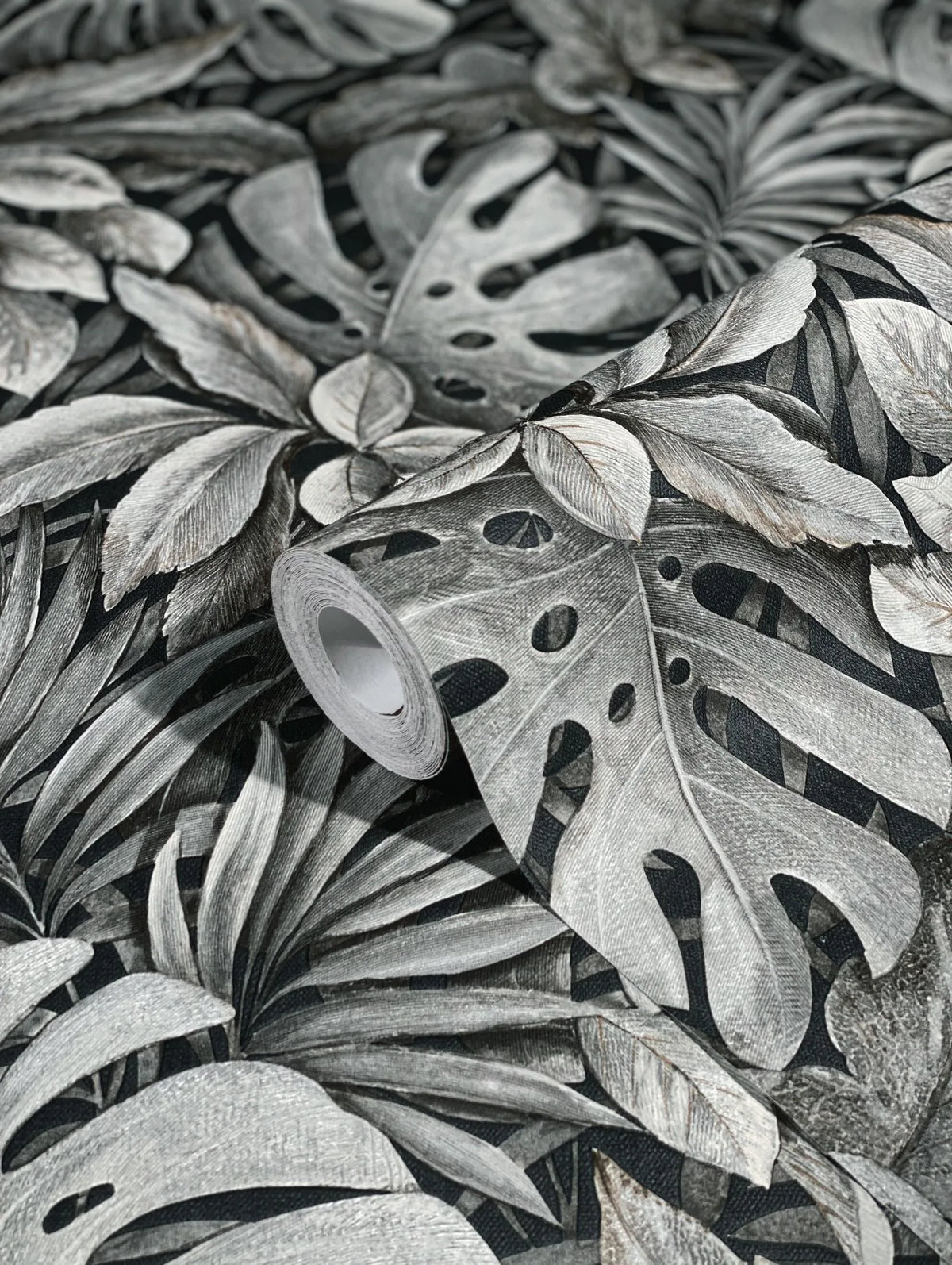Marburg Botanica 33305 A wallpaper roll resting on the same pattern, showing large overlapping tropical leaves in gray tones on a dark background.