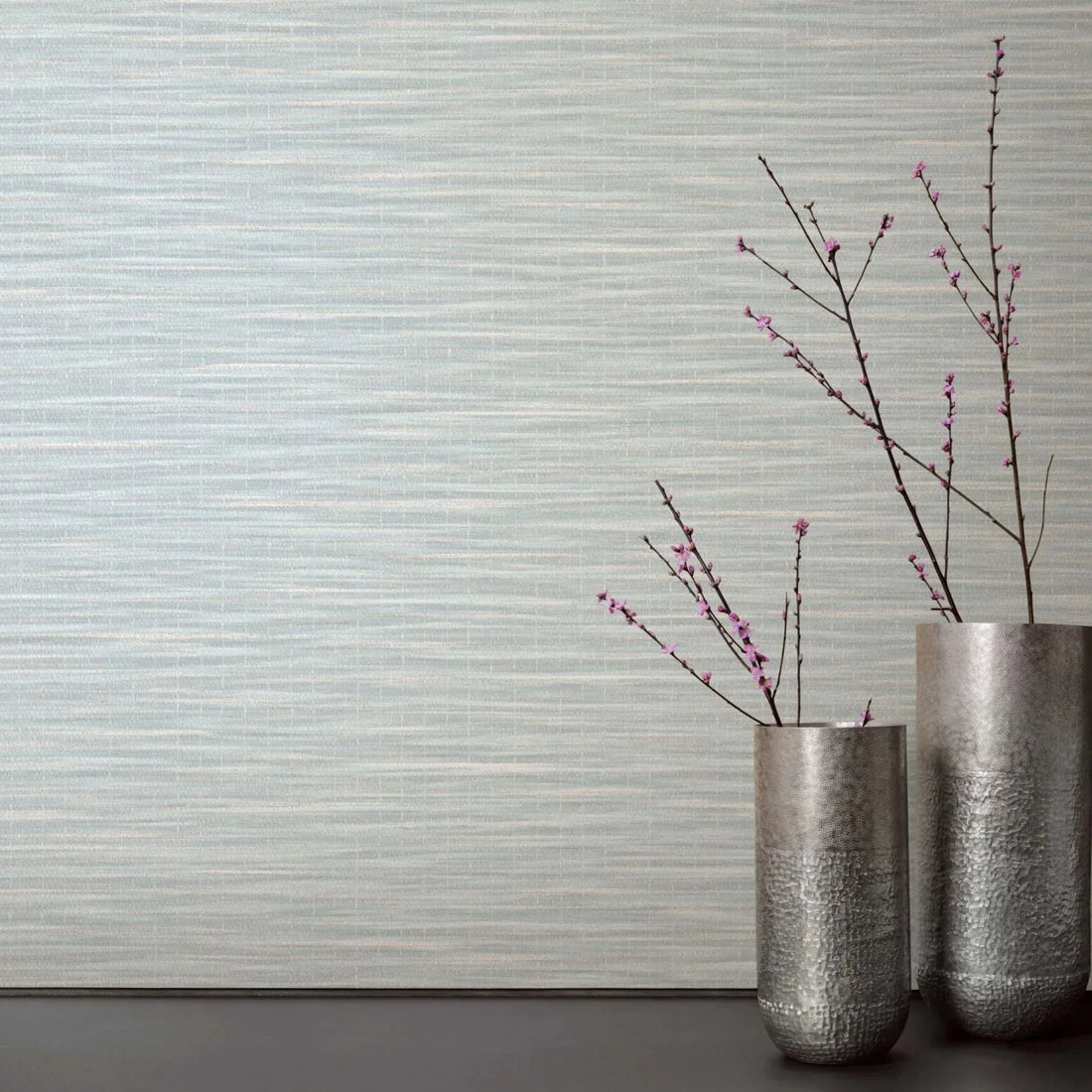 Marburg Botanica 33321 Light green and white horizontally striated wallpaper behind two tall metal vases with pink flowering branches on a dark surface.
