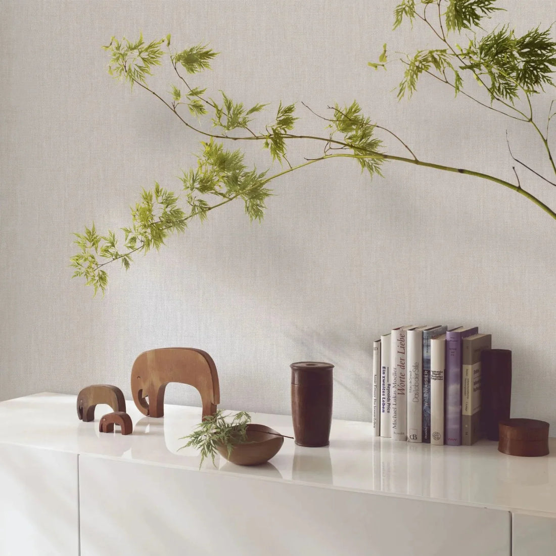 Marburg Botanica 33324 Light beige-gray textured wallpaper behind a white sideboard styled with wooden elephant figurines, a wooden bowl and cup, books, and a leafy branch.