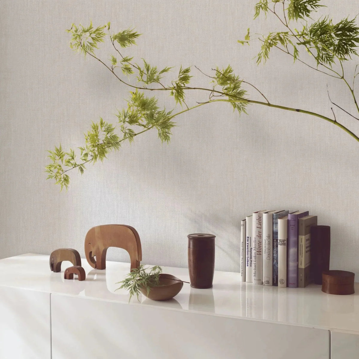 Marburg Botanica 33324 Light beige-gray textured wallpaper behind a white sideboard styled with wooden elephant figurines, a wooden bowl and cup, books, and a leafy branch.