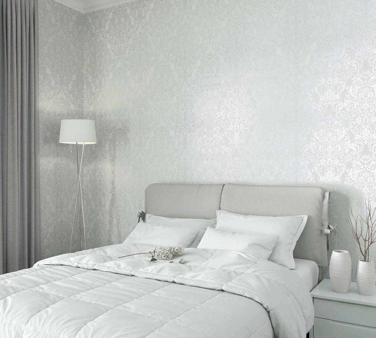 Marburg City Glam 32601 Bedroom scene featuring light gray damask wallpaper with a soft sheen behind a light upholstered headboard, white bedding, a tripod floor lamp, gray curtains, and a nightstand with two ribbed vases and twig stems.