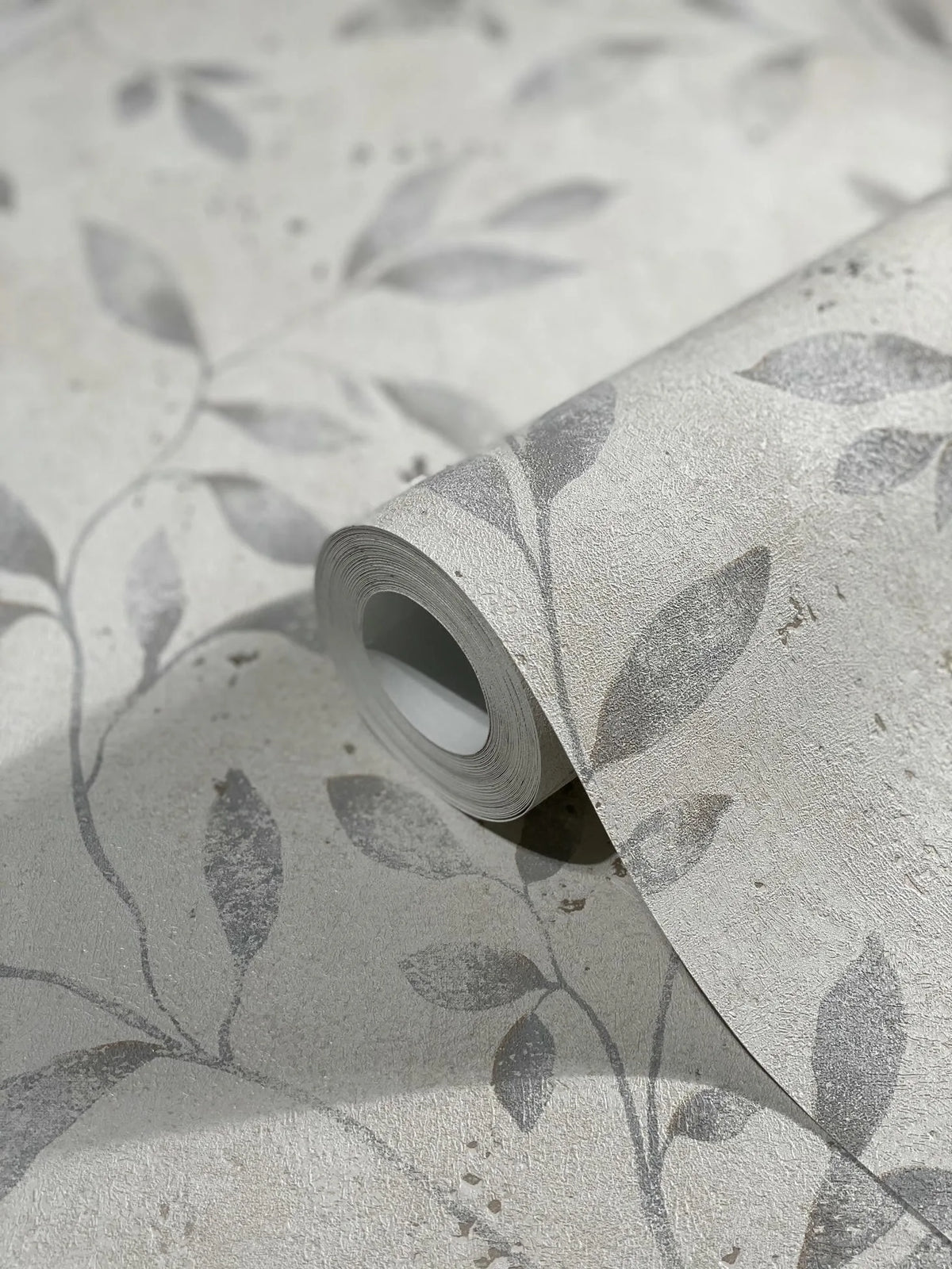 Marburg City Glam 32633 Close-up of a wallpaper roll resting on matching unrolled wallpaper, showing a textured light-neutral background with muted gray leafy vine pattern and slight speckling.