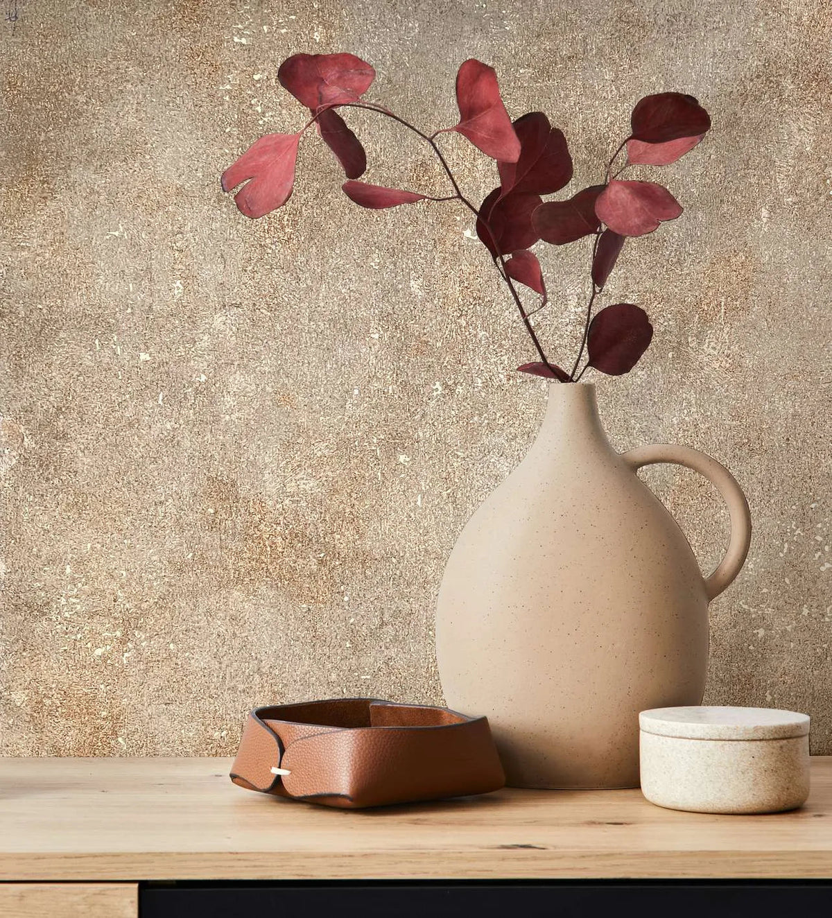 Marburg City Glow 34269 Mottled beige and brown wallpaper with a speckled texture behind a ceramic jug holding red leaves, a small leather tray, and a lidded pot on a wooden surface.