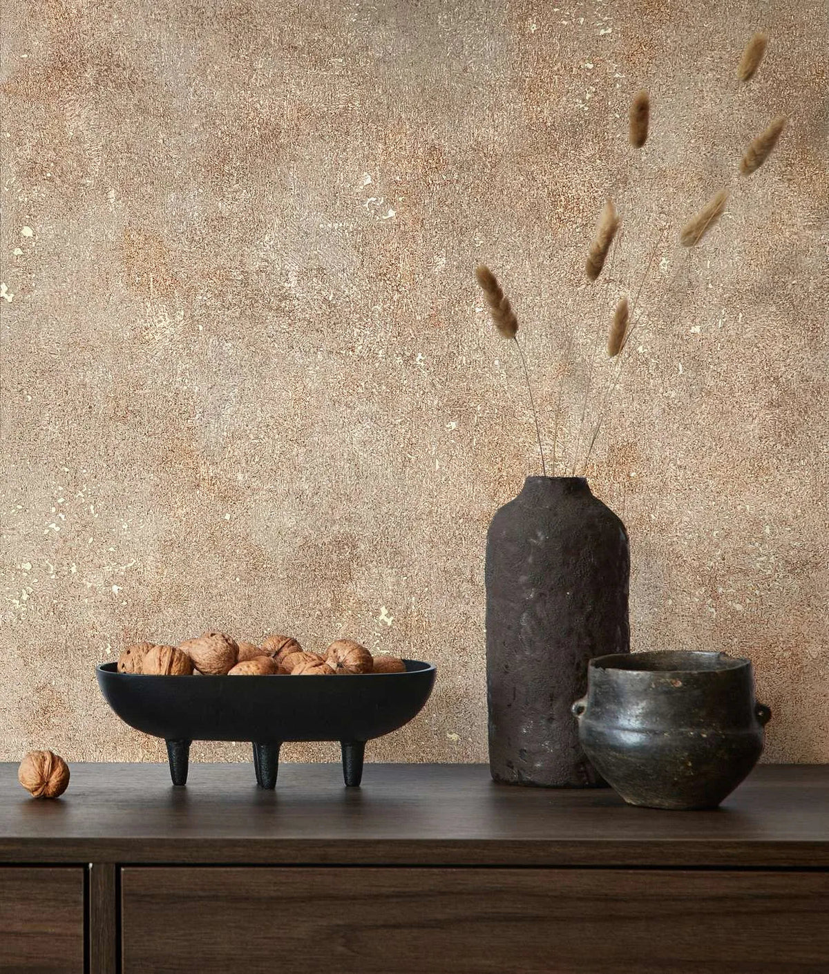 Marburg City Glow 34269 Warm beige and brown mottled wallpaper with subtle metallic flecks behind a wood sideboard holding a black tripod bowl of walnuts, a tall dark textured vase with dried stems, and a small ceramic pot.
