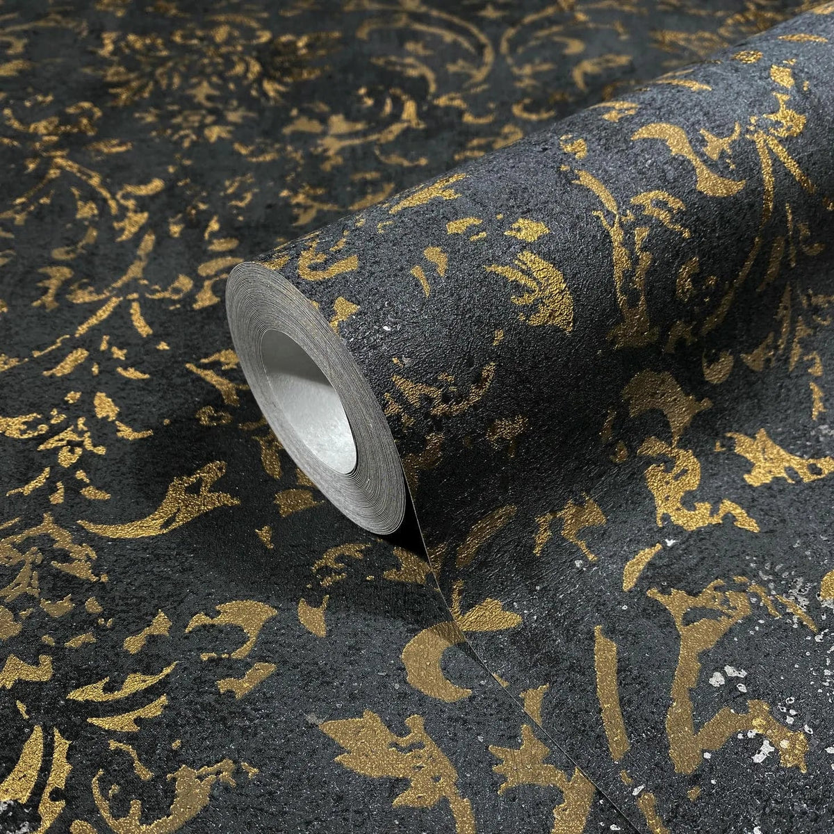 Marburg City Glow 34297 Close-up of a wallpaper roll with a metallic gold damask pattern on a dark textured background, shown partially unrolled.
