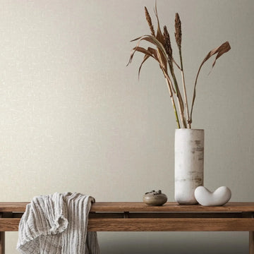Marburg City Romance 35107 Beige textured wallpaper with a subtle crosshatch pattern behind a wood bench styled with a tall white cylindrical vase holding dried stems, a draped knit throw, a small round bowl, and a small white ceramic sculpture.