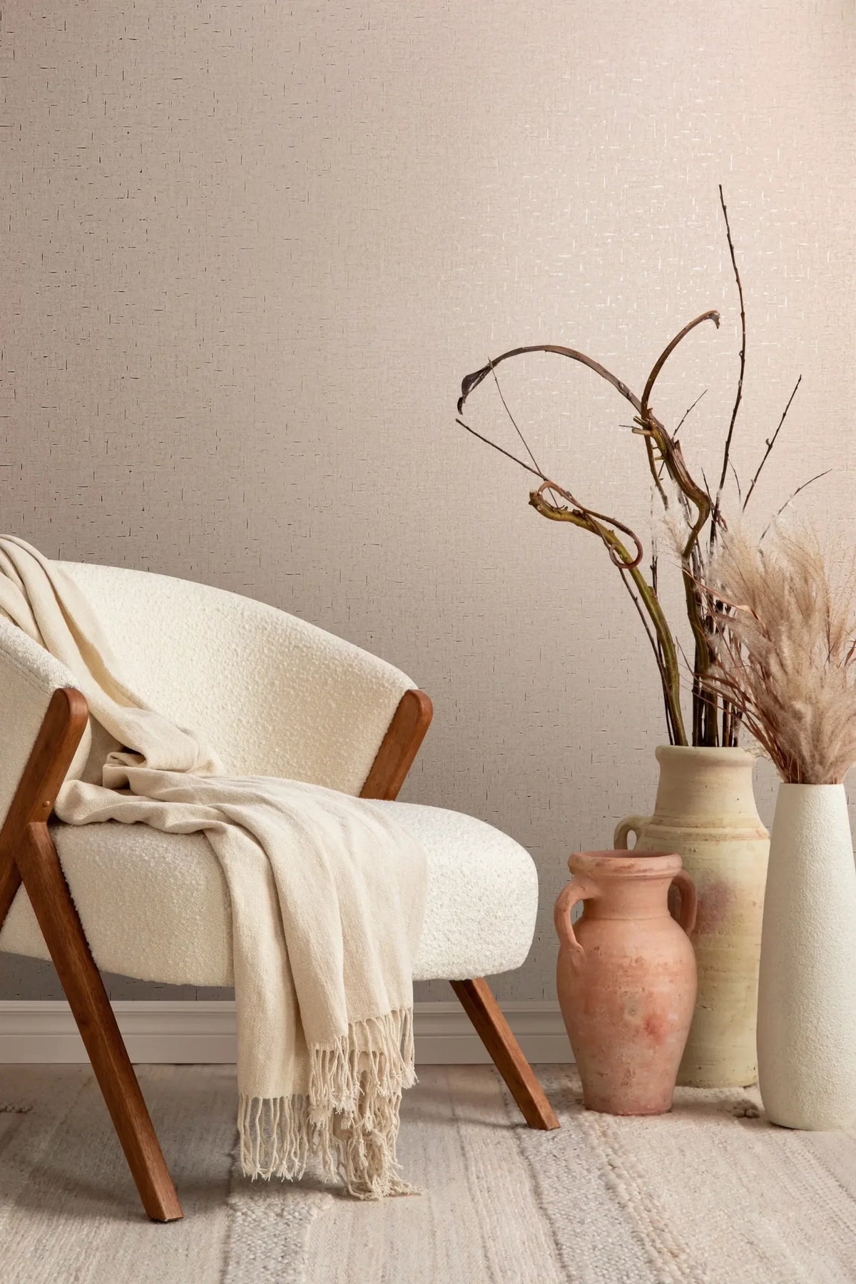 Marburg City Romance 35112 Beige wallpaper with a subtle crosshatch dash texture behind a cream armchair with a draped throw and several ceramic vases holding dried branches and pampas on a light rug.