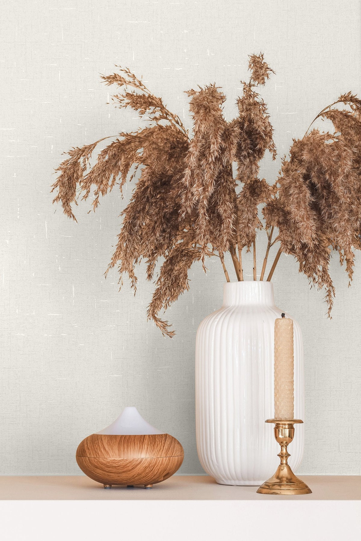 Marburg City Romance 35115 Close-up styled scene showing pale textured wallpaper with a fine crosshatch and small white slubs behind a ribbed white vase of dried pampas, a brass candlestick with a beige taper, and a wood‑grain diffuser on a shelf.