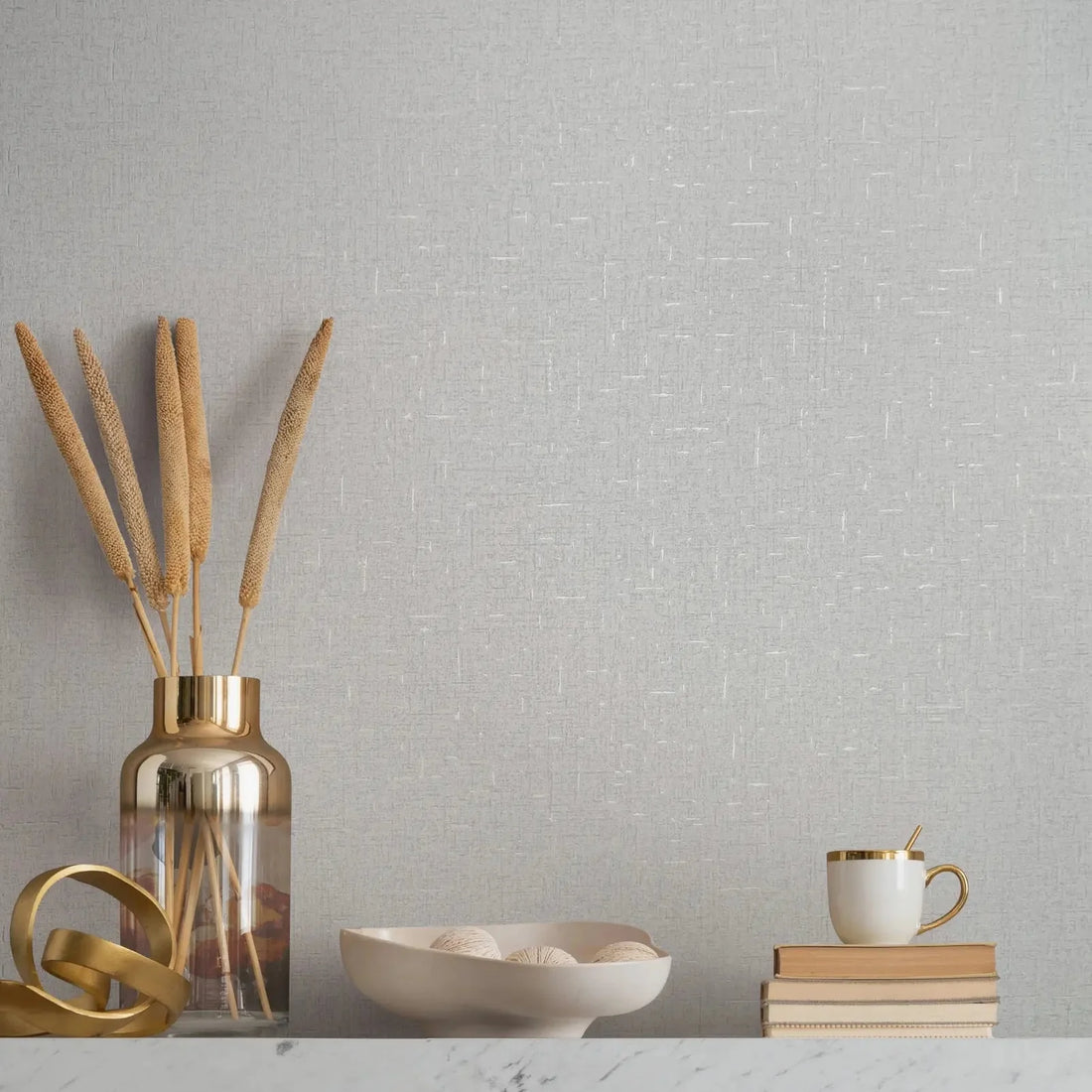 Marburg City Romance 35116 Light gray textured wallpaper with fine white crosshatch lines behind a marble-top surface styled with a metallic vase holding dried stems, a gold ribbon sculpture, a shallow bowl with decorative balls, and a stack of books topped with a mug and spoon.