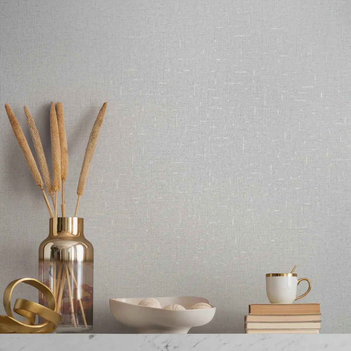 Marburg City Romance 35116 Light gray textured wallpaper with fine white crosshatch lines behind a marble-top surface styled with a metallic vase holding dried stems, a gold ribbon sculpture, a shallow bowl with decorative balls, and a stack of books topped with a mug and spoon.
