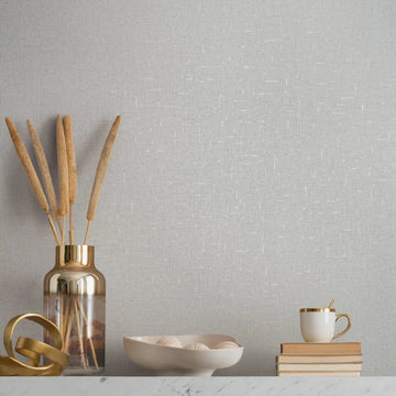 Marburg City Romance 35116 Light gray textured wallpaper with fine white crosshatch lines behind a marble-top surface styled with a metallic vase holding dried stems, a gold ribbon sculpture, a shallow bowl with decorative balls, and a stack of books topped with a mug and spoon.