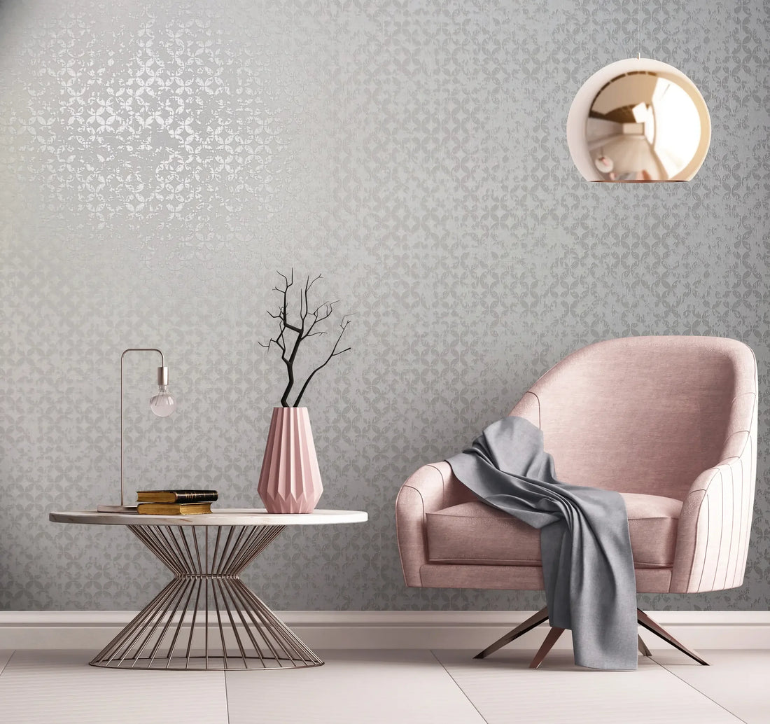 Marburg City Romance 35121 Tonal light gray damask wallpaper with a soft sheen shown in a styled room featuring a pink armchair with a draped throw, a round wire-base table with a small lamp, books, and a faceted vase with branches, plus a round pendant light.