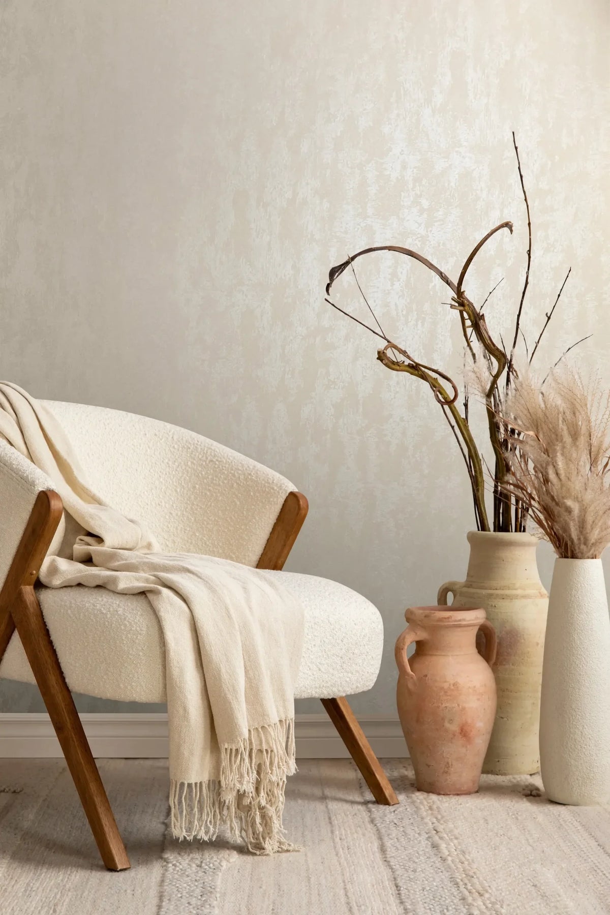 Marburg City Romance 35128 Beige textured effect wallpaper behind a cream upholstered chair with a draped throw, next to ceramic vases holding dried branches and dried grasses.