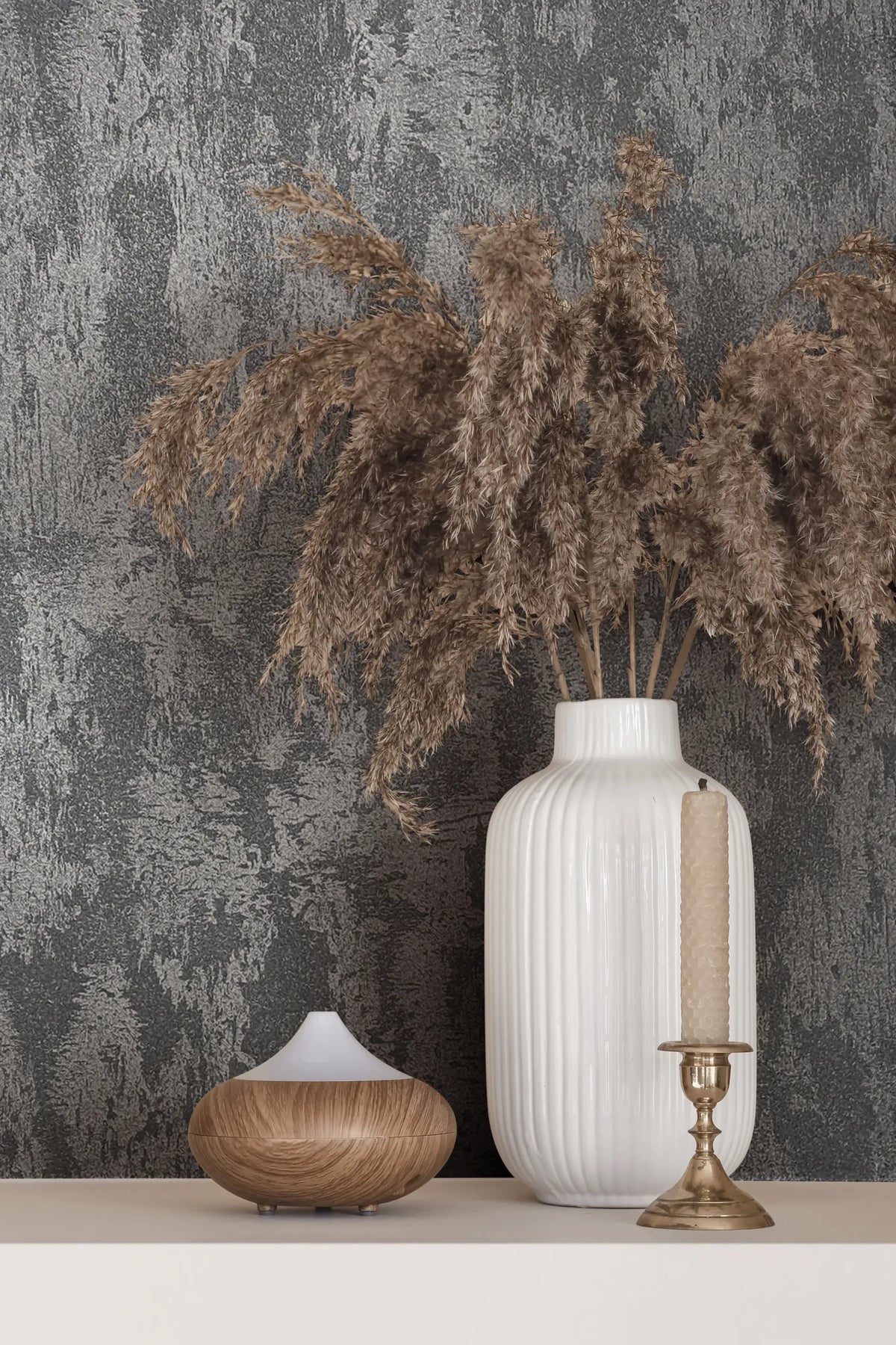Marburg City Romance 35130 Textured grey wallpaper with an abstract plaster-like pattern behind a white ribbed vase holding dried grasses, a brass candlestick with a tapered candle, and a small wood-grain diffuser on a light shelf.