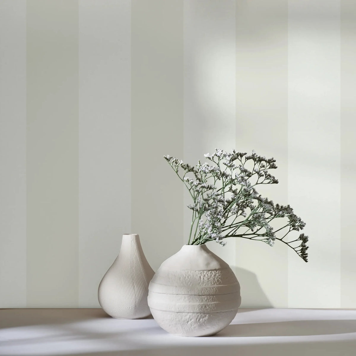 Marburg 47633 wallpaper lounge feature wall with wide vertical stripes behind a simple tabletop display of two white ceramic vases, one with delicate sprigs, serene and minimalist.