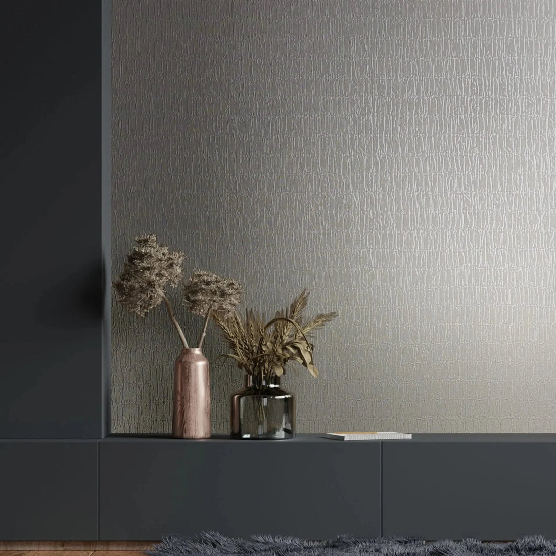 Marburg Kumano 34503 Gray wallpaper with a subtle irregular linear texture shown as a feature wall behind a dark cabinet, with two vases holding dried stems, a book, a shag rug, and a wood floor in view.