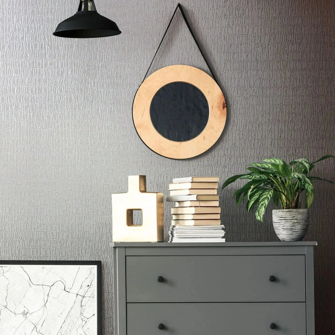 Marburg Kumano 34506 Room vignette showing gray textured wallpaper with an irregular vertical pattern behind a gray three-drawer dresser. A round hanging mirror and a black pendant light are on the wall, with a potted plant, stacked books, and a geometric sculpture on the dresser.