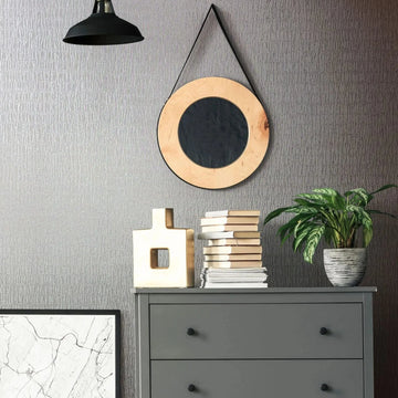 Marburg Kumano 34506 Room vignette showing gray textured wallpaper with an irregular vertical pattern behind a gray three-drawer dresser. A round hanging mirror and a black pendant light are on the wall, with a potted plant, stacked books, and a geometric sculpture on the dresser.