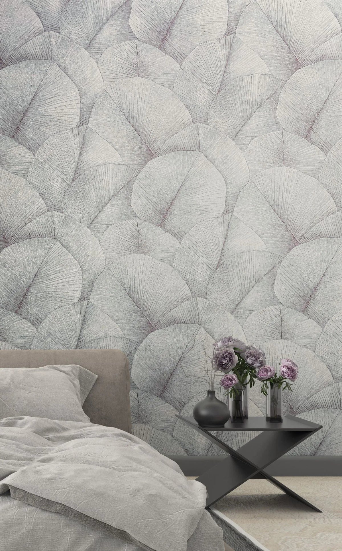 Marburg Kumano 34512 Bedroom scene featuring a large-scale leaf pattern wallpaper in light gray behind a bed with gray bedding and a black side table holding vases with purple flowers.