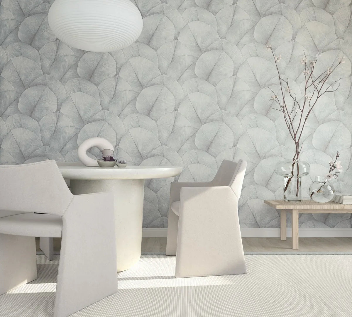 Marburg Kumano 34512 Room scene with a wall covered in soft gray overlapping fan‑leaf wallpaper behind a round light-toned dining table, sculptural armchairs, a pendant light, and a wooden bench holding clear glass vases with branches.