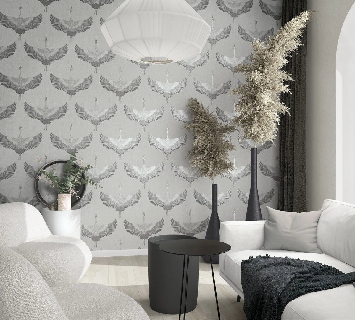 Marburg Kumano 34530 Gray wallpaper with repeating cranes in flight shown in a living room setting with a white sofa and lounge chair, black round nesting tables, tall black floor vases with feathery branches, a white pendant light, and an arched doorway.