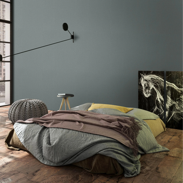 Marburg Loft Superior 34181 Textured green wallpaper on the feature wall behind a low bed in a modern bedroom, with a long-arm wall sconce, knitted pouf, small stool table, large grid window, and horse artwork panels leaning against the wall.