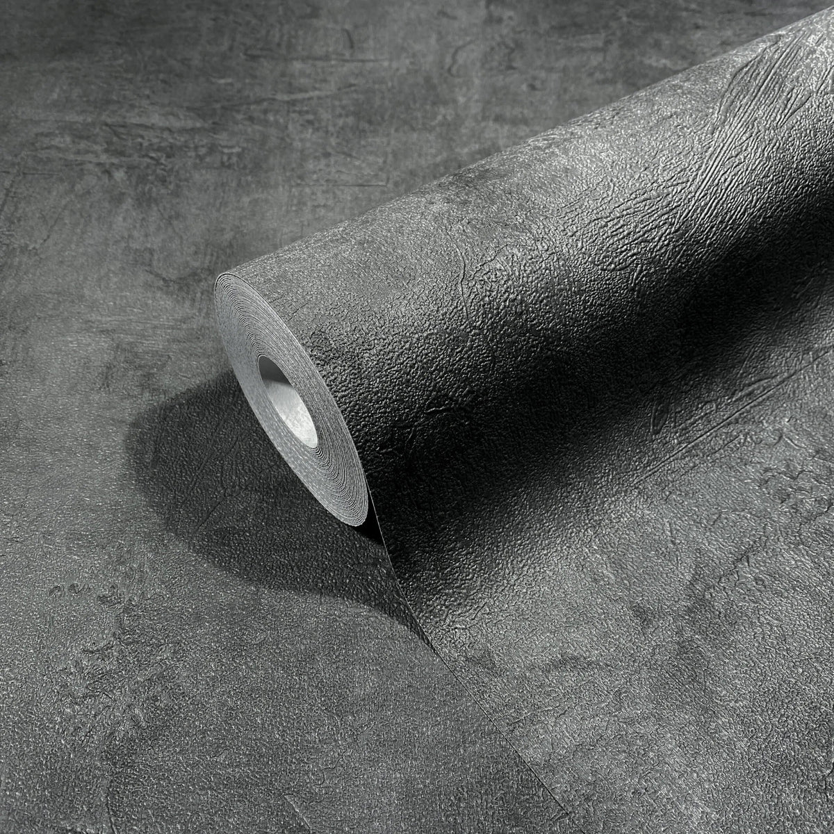 Marburg Loft Superior 34193 A dark anthracite wallpaper roll partially unrolled, displaying a textured, plaster-like surface.