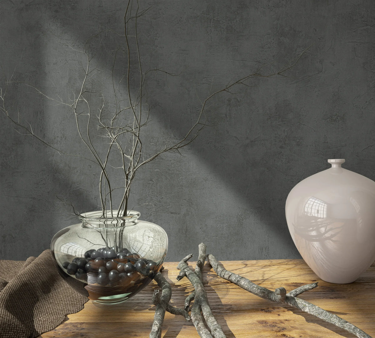 Marburg Loft Superior 34193 Dark grey concrete-look wallpaper with a mottled texture behind a wood tabletop styled with a clear glass vase of branches, dark pebbles, a light ceramic jar, loose twigs, and a brown cloth.