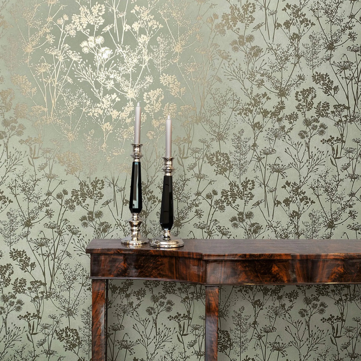 Marburg Mona 82465 Green wallpaper with metallic gold and dark green botanical silhouettes behind a dark wood console table with two tall candlesticks and white taper candles.