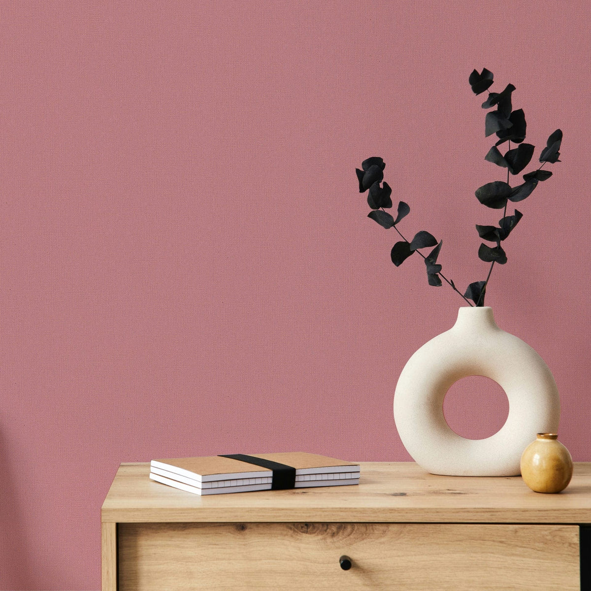 Marburg Mona 82467 Solid pink textured wallpaper behind a light wood cabinet topped with a round cutout ceramic vase holding dark foliage, a small round vase, and stacked notebooks.