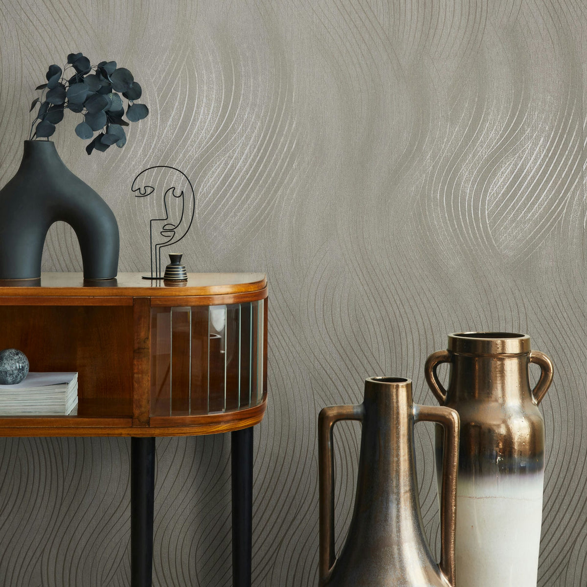 Marburg Mona 82475 Beige-gray wallpaper with shimmering wavy lines shown with a wood console, a black sculptural vase with foliage, a wire face ornament, and two tall metallic vases.