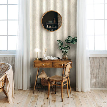 Marburg Mona 89006 Room scene featuring light beige vertical ribbed stripe wallpaper behind a wood desk and chair, with a round mirror, sheer curtains over large windows, a table lamp, a tray with a teapot and cups, and a leafy plant in a glass vase.