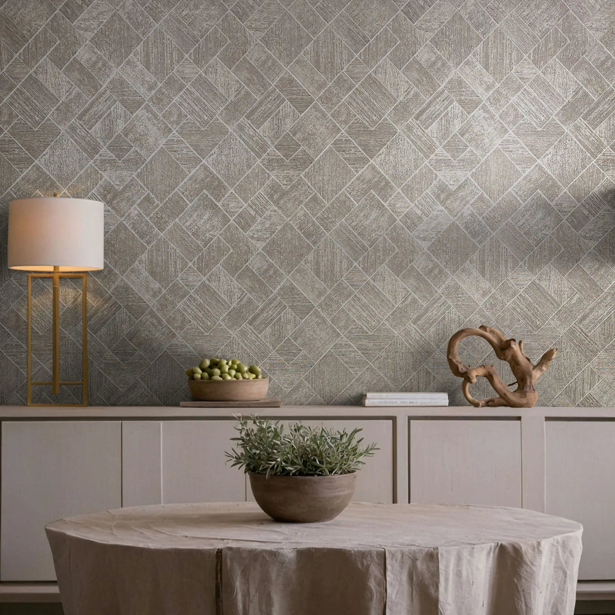 Marburg Ritus 35510 Beige and gray geometric diamond-pattern wallpaper behind a styled dining vignette with a round draped table holding a potted plant, a sideboard with a lamp, books, a wooden sculpture, and a bowl of green fruit.