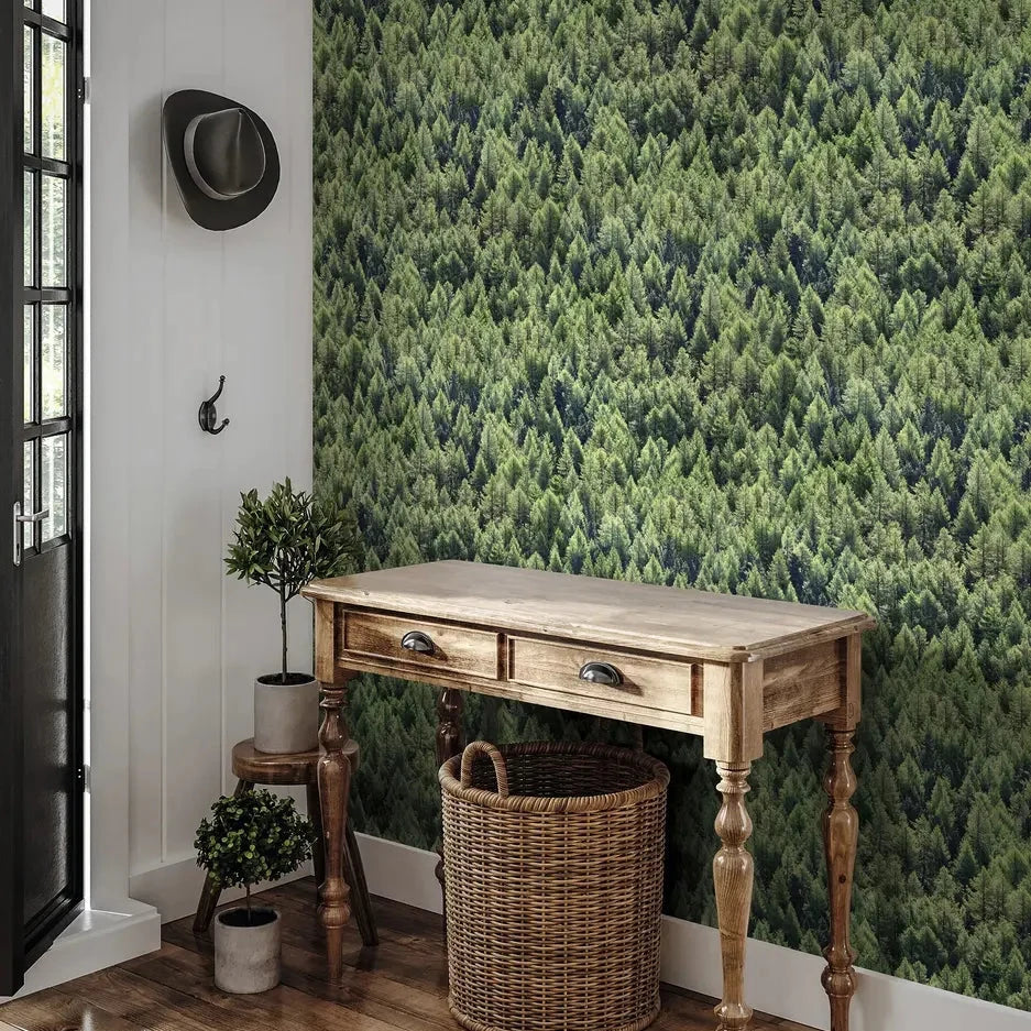 Marburg Struktura 45730 Green forest treetops wallpaper behind a wooden console table with a wicker basket; potted plants, a wall hook with a hat, and a door are visible.