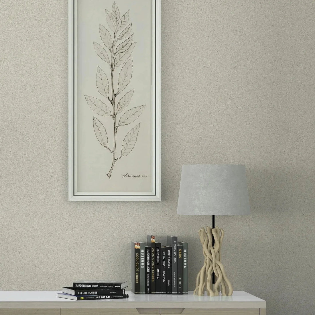 Marburg Travertino 33363 Light beige textured wallpaper behind a modern sideboard styled with stacked books, a sculptural table lamp, and a tall framed leaf illustration.