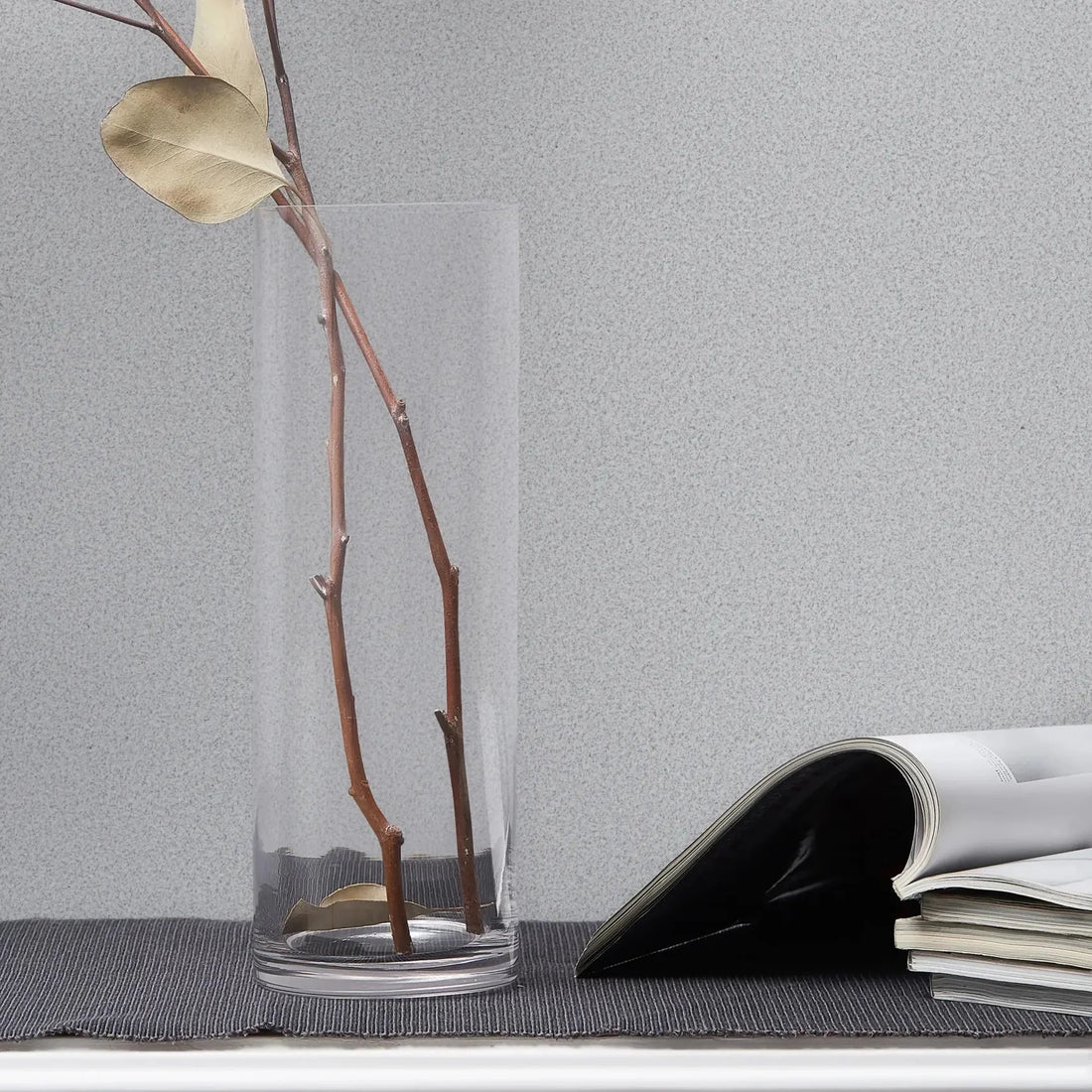 Marburg Travertino 33365 Light gray speckled wallpaper shown behind a clear glass cylinder vase with bare branches and an open magazine on a white molded console.