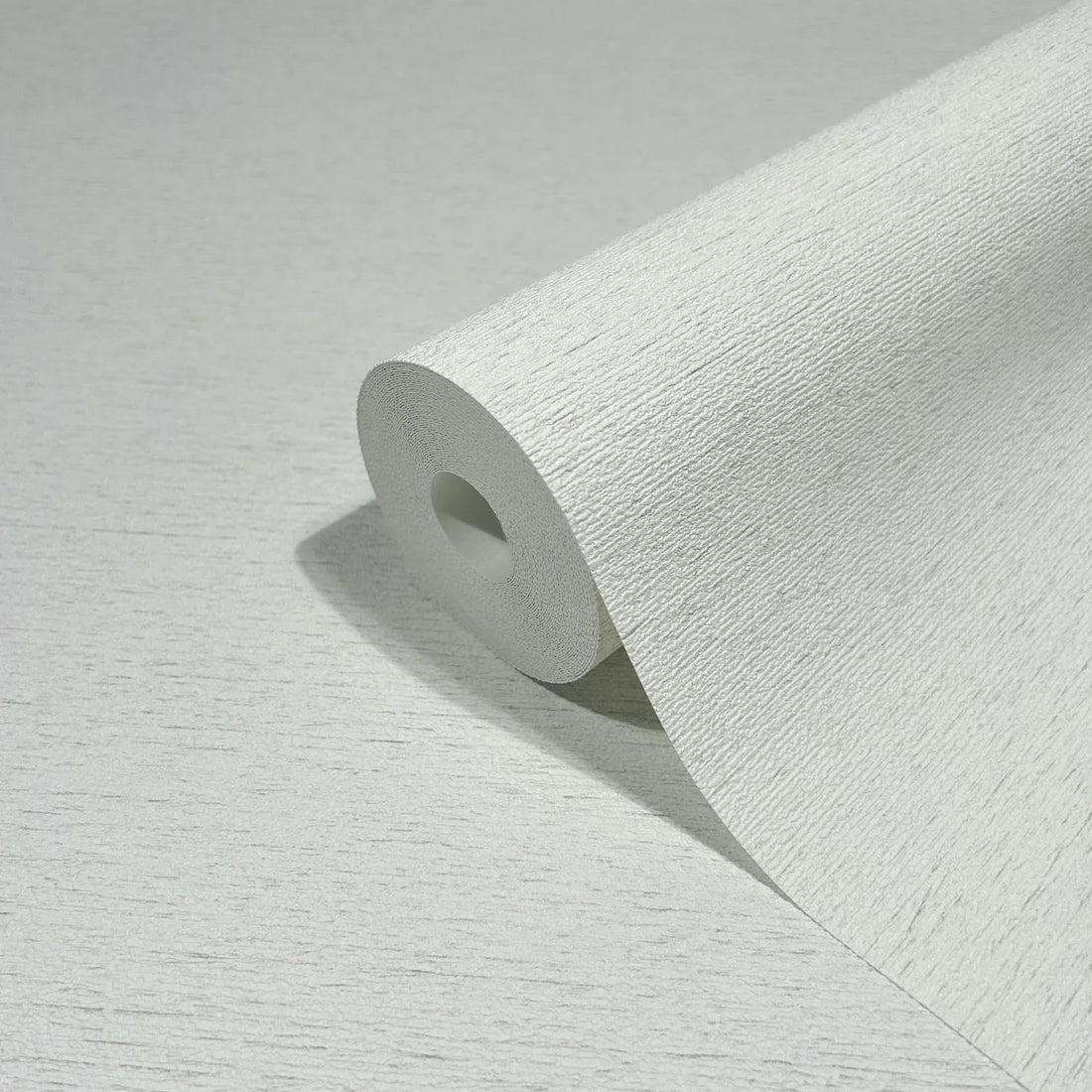 Marburg Travertino 33368 Light textured off-white wallpaper behind a styled bedroom scene with a bed, a small white bedside table topped with a ribbed glass vase and greenery, a floor vase with eucalyptus, and a white shelving unit.