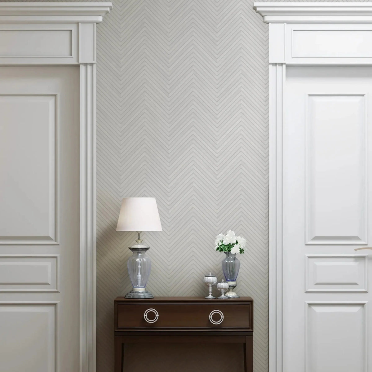 Marburg Travertino 33375 Light gray chevron wallpaper behind a dark wood console table set between two white paneled doors. The table holds a glass-base lamp, a small vase with white flowers, and decorative jars on a wood floor.