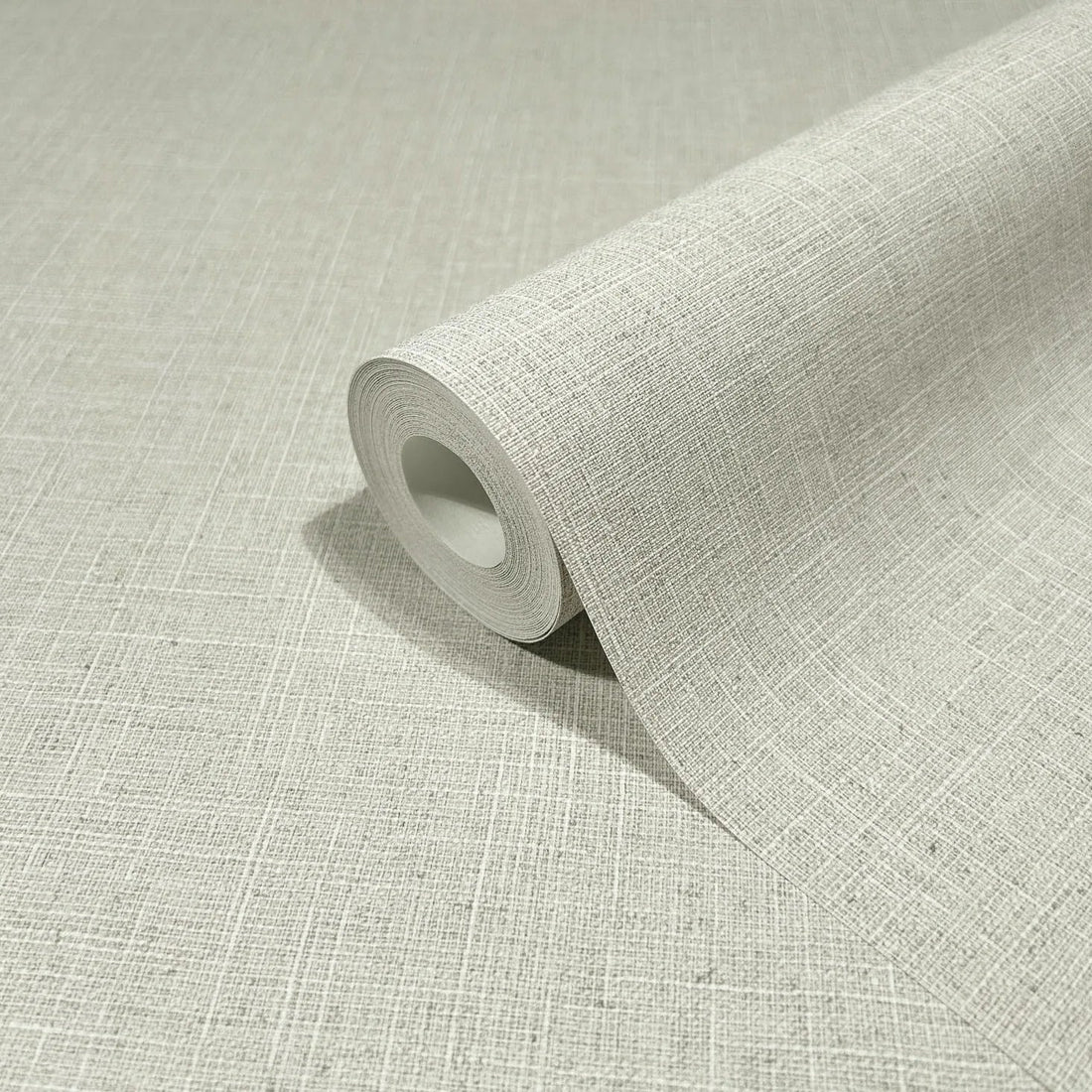 Marburg Travertino 33388 Light beige-gray linen-look textured wallpaper behind a boucle chair, a floor planter of dried grasses, and decorative pillows on a wood floor with baseboard trim.